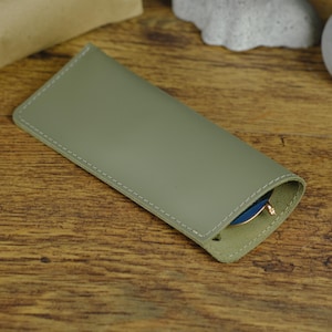 May include: A green leather glasses case with a stitched edge. The case is closed and a pair of glasses is visible inside.