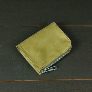以下が含まれることがあります： 滑らかでマットな質感のオリーブグリーンのレザー製小型財布。縁にはステッチが施されています。財布の上部から側面にかけてシルバーカラーの金属製ファスナーがカーブを描いて取り付けられており、コンパクトなアクセサリーをしっかりと閉じることができます。
