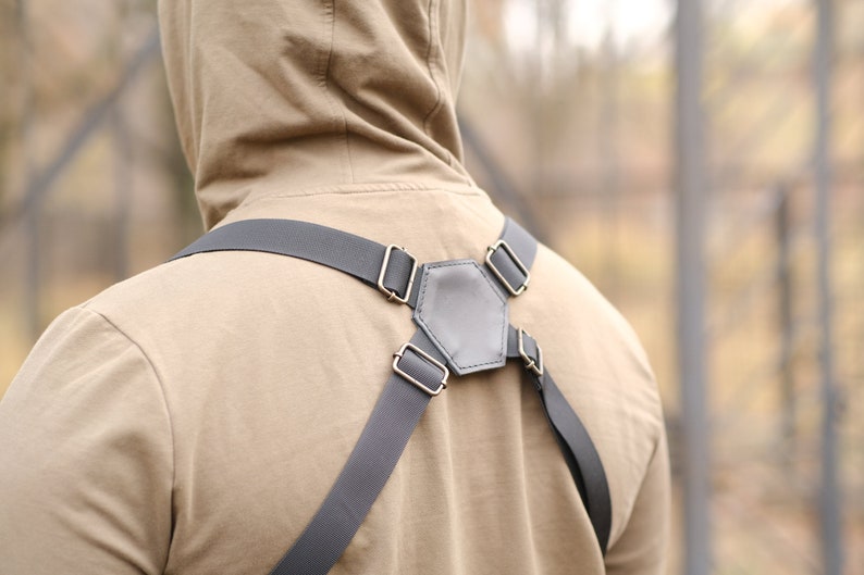 May include: A dark gray harness featuring adjustable elastic straps and a central hexagonal leather-like back panel. The harness includes metallic rectangular sliders and rings for adjustment, connecting the straps in a symmetrical configuration.