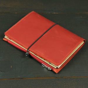 Puede incluir: Una funda de cuaderno de cuero rojo con acabado mate y grano visible. La funda está asegurada por un cordón elástico negro fino envuelto verticalmente alrededor del centro. Un pequeño bolsillo rojo con cremallera está unido al borde inferior con un tirador de metal plateado.