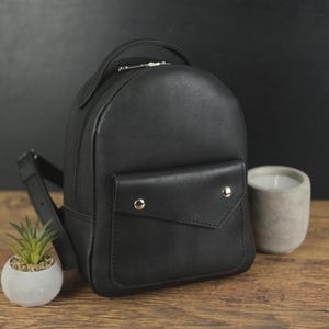 Puede incluir: Una mochila negra con acabado liso, que cuenta con un asa superior, correas ajustables para los hombros y un bolsillo frontal con solapa asegurado por dos botones de presión de metal plateado. El diseño presenta una parte superior redondeada.