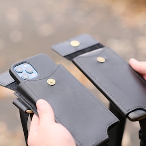 May include: Two black leather phone pouches with a flap closure secured by a brass-colored metal snap button. One pouch contains a smartphone with a visible triple-lens camera array. Each pouch features a matching black strap for carrying.