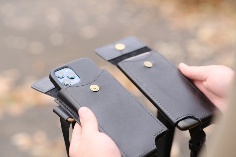 May include: Two black leather phone pouches with a flap closure secured by a brass-colored metal snap button. One pouch contains a smartphone with a visible triple-lens camera array. Each pouch features a matching black strap for carrying.