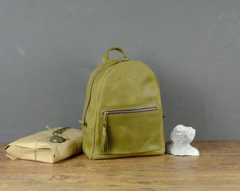 Può includere: Uno zaino in pelle verde oliva con manico superiore, uno scomparto principale con cerniera e una tasca frontale con cerniera e tirante con nappa in pelle abbinata. Accanto allo zaino si trova un pacchetto rettangolare in carta marrone e un busto bianco.