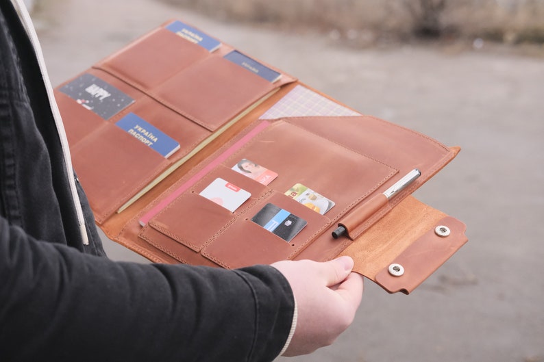 Puede incluir: Un organizador de viaje de cuero color canela con m&uacute;ltiples ranuras interiores para pasaportes, tarjetas de cr&eacute;dito y documentos de identidad. La cartera incluye un soporte para bol&iacute;grafo en el lado derecho y dos broches met&aacute;licos plateados.