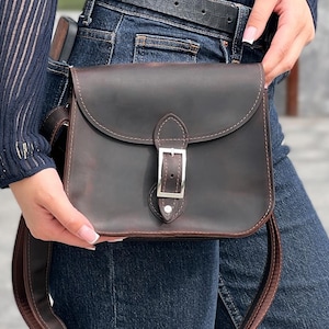 May include: A dark brown leather satchel-style crossbody bag with a flap and silver buckle. The bag has an adjustable strap. The bag is made of leather and is worn over the shoulder. The bag is a classic design.