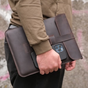 以下が含まれることがあります： フラップ留めとストラップが付いた茶色の革製ラップトップスリーブ。スリーブには、電話やその他の小物を入れるためのフロントポケットがあります。スリーブは人が持っています。