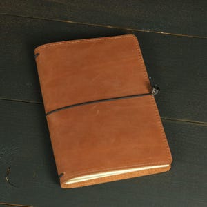 May include: A rectangular notebook with a tan leather cover featuring a smooth, matte texture. A thin black elastic band wraps horizontally around the center of the cover, secured by a small, dark metallic bead detail on the right side.