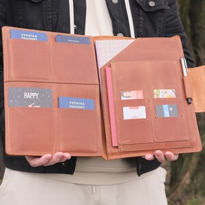 Puede incluir: Un organizador de documentos de cuero color canela con cuatro ranuras para pasaportes, varios bolsillos para tarjetas, un compartimento para cuaderno y un portal&aacute;pices. El interior contiene dos pasaportes azules UKRAINA PASPORT y varias tarjetas.