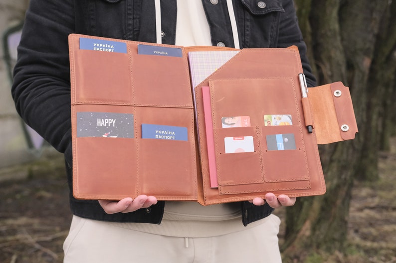 Puede incluir: Un organizador de documentos de cuero color canela con cuatro ranuras para pasaportes, varios bolsillos para tarjetas, un compartimento para cuaderno y un portal&aacute;pices. El interior contiene dos pasaportes azules UKRAINA PASPORT y varias tarjetas.