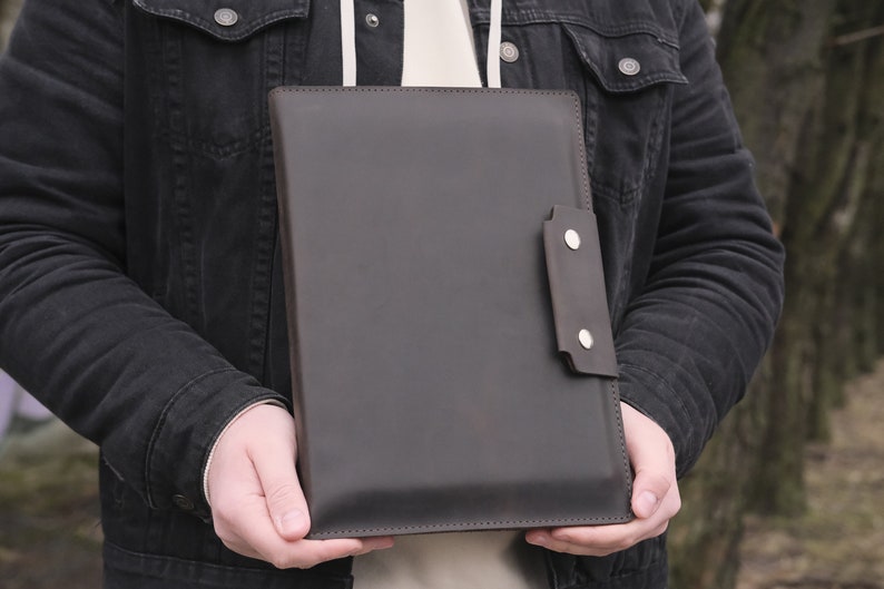 Puede incluir: Una carpeta para documentos de cuero marr&oacute;n oscuro con forma rectangular, superficies lisas y un cierre de solapa lateral asegurado con dos botones de presi&oacute;n de metal plateado. La carpeta tiene costuras visibles a lo largo de los bordes.