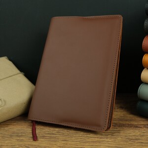 May include: A brown leather bound journal with a red ribbon bookmark. The journal is lying on a wooden surface.