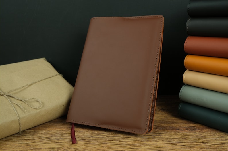 May include: A brown leather bound journal with a red ribbon bookmark. The journal is lying on a wooden surface.