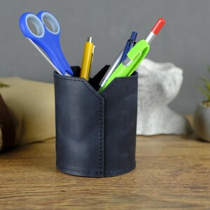 Peut inclure: Un porte-crayon cylindrique en cuir bleu marine foncé avec des coutures verticales visibles sur le côté. Le support contient une paire de ciseaux à poignées bleues, un stylo jaune, un stylo argenté et un stylo vert citron vif avec un capuchon rouge.