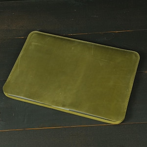 May include: A rectangular olive green leather laptop sleeve with rounded corners and visible stitching along the edges. The surface of the sleeve has a smooth, matte texture with subtle natural variations and light scuff marks across the front.