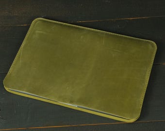 May include: A rectangular olive green leather laptop sleeve with rounded corners and visible tonal stitching along the edges. The surface of the sleeve has a smooth, matte texture with subtle natural variations and light scuff marks throughout.