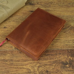 May include: A brown leather bound journal with a red ribbon bookmark. The journal is lying on a wooden surface.
