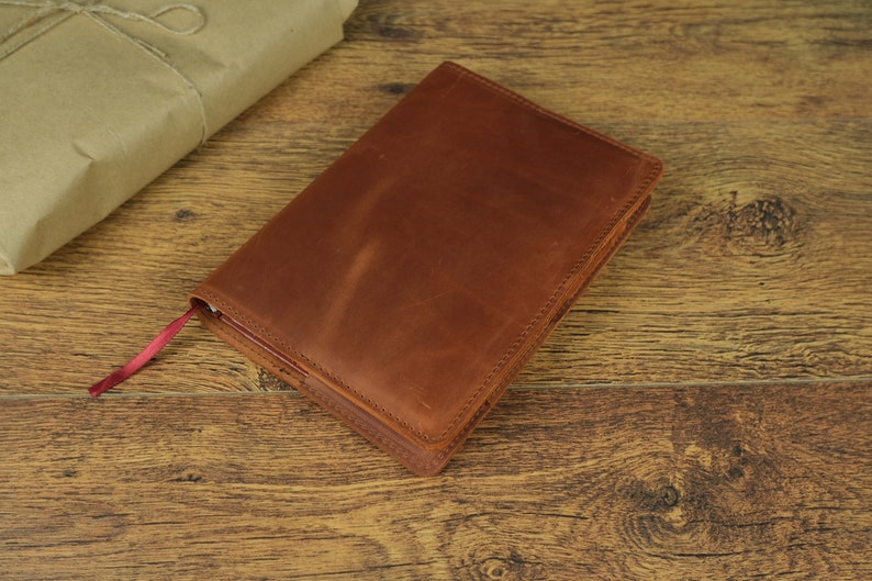 May include: A brown leather bound journal with a red ribbon bookmark. The journal is lying on a wooden surface.