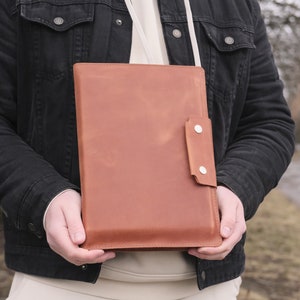 Puede incluir: Un portadocumentos rectangular de cuero color canela con un cierre de solapa vertical asegurado por dos botones de presi&oacute;n de metal plateado. El estuche de cuero liso con acabado mate incluye una correa fina de color claro.
