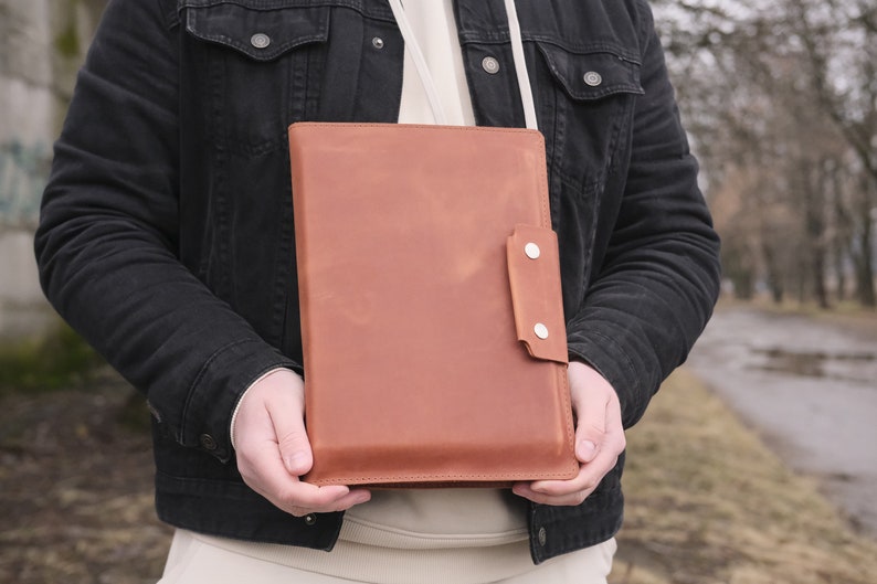 Puede incluir: Un portadocumentos rectangular de cuero color canela con un cierre de solapa vertical asegurado por dos botones de presi&oacute;n de metal plateado. El estuche de cuero liso con acabado mate incluye una correa fina de color claro.