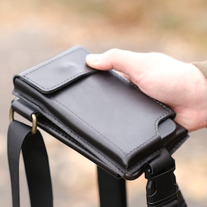 May include: A black leather phone holster with a flap closure and a matching black nylon strap. The holster features visible stitching along the edges and is attached to a black plastic buckle and a metallic ring for secure carrying and attachment.