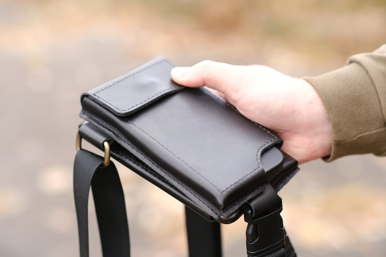 May include: A black leather phone holster with a flap closure and a matching black nylon strap. The holster features visible stitching along the edges and is attached to a black plastic buckle and a metallic ring for secure carrying and attachment.