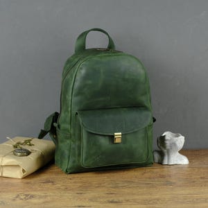 Puede incluir: Una mochila de cuero verde con un bolsillo frontal y un cierre dorado. Un paquete envuelto con un colgante de estilo vintage y una pequeña escultura gris están junto a la mochila. Los artículos están sobre una superficie de madera.