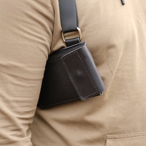 May include: A dark brown leather holster-style pouch featuring a rectangular flap closure with visible stitching. The pouch is attached to a black adjustable nylon strap that rests over the shoulder, secured by a metallic D-ring hardware component.