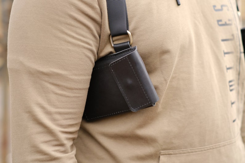 May include: A dark brown leather holster-style pouch featuring a rectangular flap closure with visible stitching. The pouch is attached to a black adjustable nylon strap that rests over the shoulder, secured by a metallic D-ring hardware component.