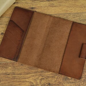 May include: Open brown leather notebook with a flap and a pocket on the front cover. The notebook is lying open on a wooden surface.