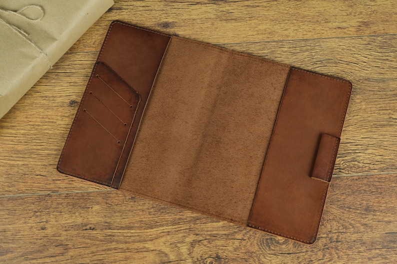 May include: Open brown leather notebook with a flap and a pocket on the front cover. The notebook is lying open on a wooden surface.