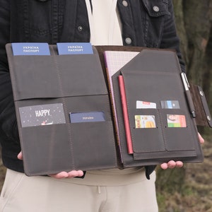 Puede incluir: Un organizador de viaje de cuero marr&oacute;n oscuro con m&uacute;ltiples ranuras interiores para documentos, incluyendo dos pasaportes ucranianos azules, una tarjeta con la palabra HAPPY, un cuaderno peque&ntilde;o, cuatro tarjetas de cr&eacute;dito y un bol&iacute;grafo plateado.