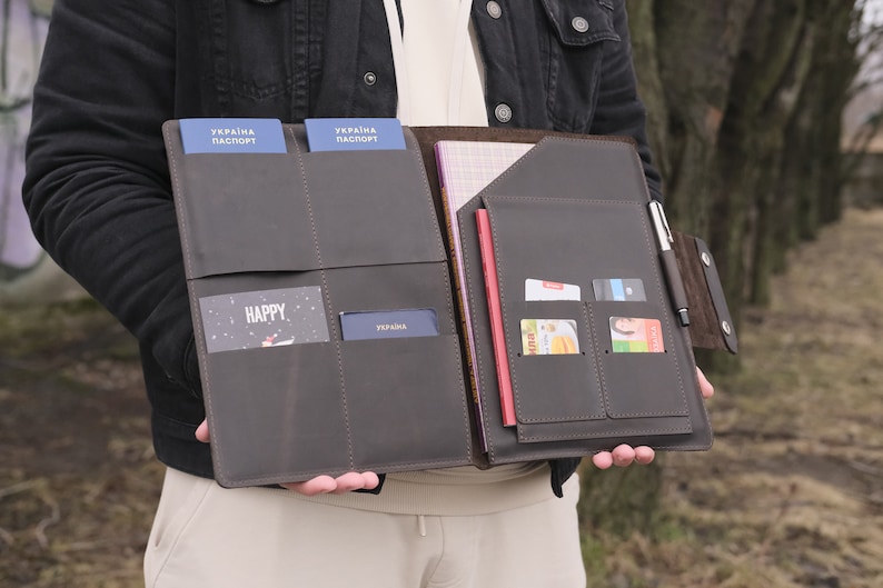 Puede incluir: Un organizador de viaje de cuero marr&oacute;n oscuro con m&uacute;ltiples ranuras interiores para documentos, incluyendo dos pasaportes ucranianos azules, una tarjeta con la palabra HAPPY, un cuaderno peque&ntilde;o, cuatro tarjetas de cr&eacute;dito y un bol&iacute;grafo plateado.