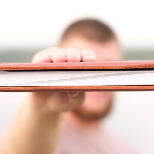 May include: A brown leather-bound journal with white pages. The journal is being held in front of a person's face.