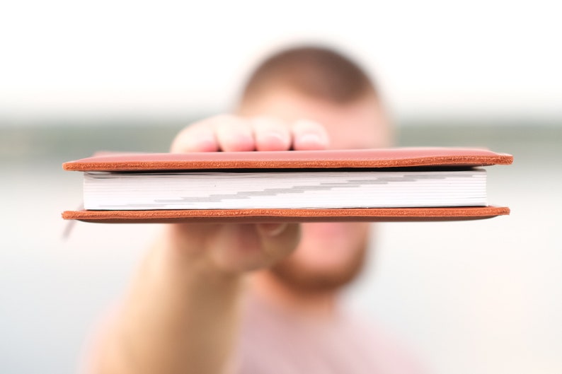 May include: A brown leather-bound journal with white pages. The journal is being held in front of a person's face.