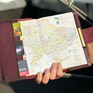 May include: A burgundy leather-bound journal with a map of Ukraine inside. The map is open to show the country's borders and major cities. The journal has a pen holder and a pocket for cards.