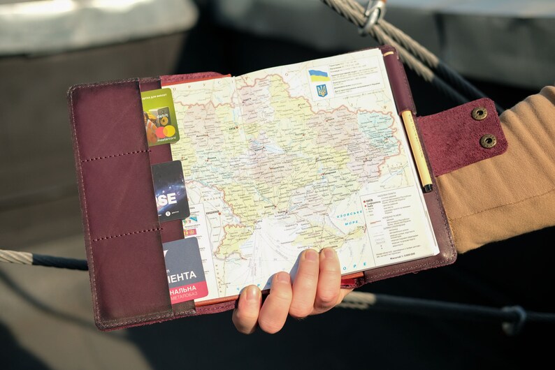 May include: A burgundy leather-bound journal with a map of Ukraine inside. The map is open to show the country's borders and major cities. The journal has a pen holder and a pocket for cards.