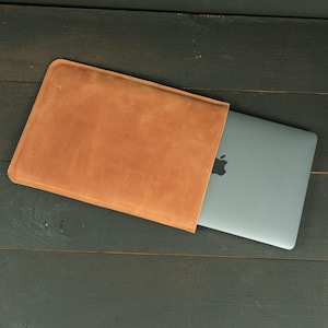 May include: A tan leather laptop sleeve with rounded corners, partially containing a silver laptop with a visible black apple logo on the lid. The sleeve has a smooth, matte texture and stitched edges, providing a protective cover for the device.