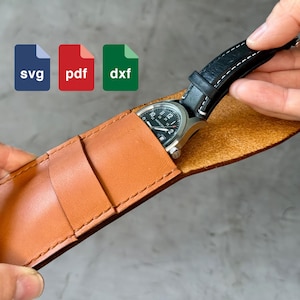 Könnte beinhalten: Ein braunes Lederuhrenetui mit einer schwarzen Uhr mit weißem Zifferblatt und schwarzen Zeigern im Inneren. Das Uhretui hat eine Klappe, die sich über die Uhr klappen lässt. Das Uhretui wird von einer Hand gehalten.