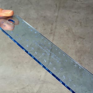 May include: A clear plastic ruler with blue edges and markings. The ruler is 12 inches long and has markings for inches and fractions of an inch. The ruler is labeled "Sewing Rule".