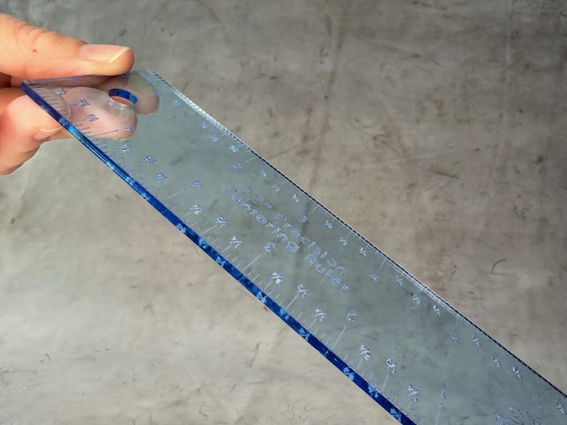 May include: A clear plastic ruler with blue edges and markings. The ruler is 12 inches long and has markings for inches and fractions of an inch. The ruler is labeled "Sewing Rule".