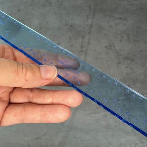 May include: A clear blue plastic ruler with black markings. The ruler is 12 inches long and has markings for every 1/16th of an inch.