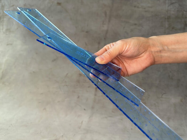 May include: Three blue plastic rulers stacked together. The rulers are marked with measurements in inches.