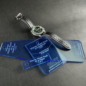 May include: Three blue plastic templates for a watch pouch, labeled "Warren Watch Pouch 4.5 oz. Outside Panel 1 of 3", "Warren Watch Pouch 4.5 oz. Pocket Panel 2 of 3", and "Warren Watch Pouch 4.5 oz. Keeper 3 of 3". The templates are on a gray surface with a black watch with a green face.