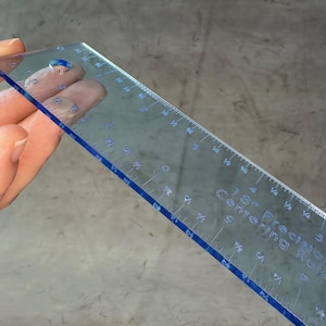 May include: A clear plastic ruler with blue edges and markings. The ruler is 18 inches long and has markings for 1/8 inch increments. The ruler is labeled "Precision 18" Centering Ruler".