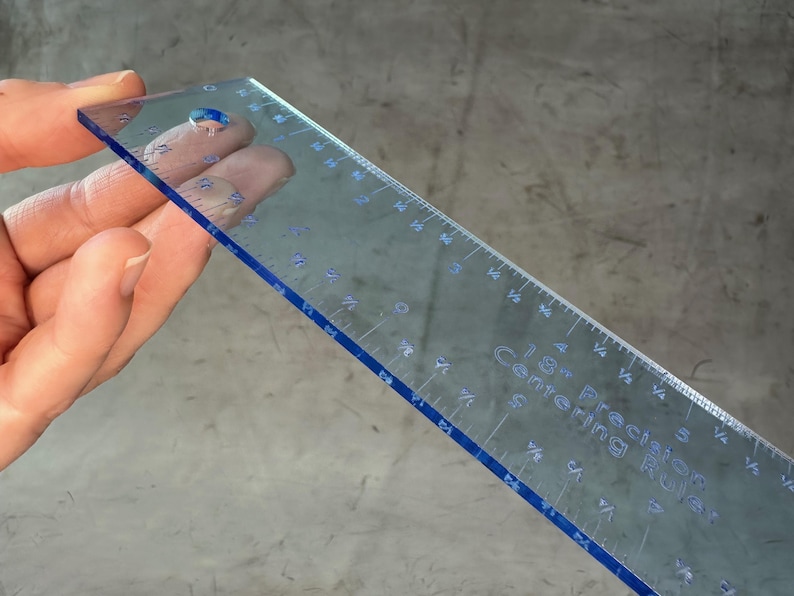 May include: A clear plastic ruler with blue edges and markings. The ruler is 18 inches long and has markings for 1/8 inch increments. The ruler is labeled "Precision 18" Centering Ruler".