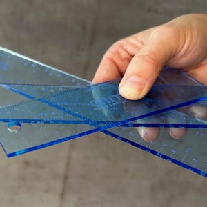May include: Two blue plastic rulers with a clear background. The rulers are marked with inches and fractions of an inch. The top ruler has the text "GRITT" and "MENSANTILE" printed on it.