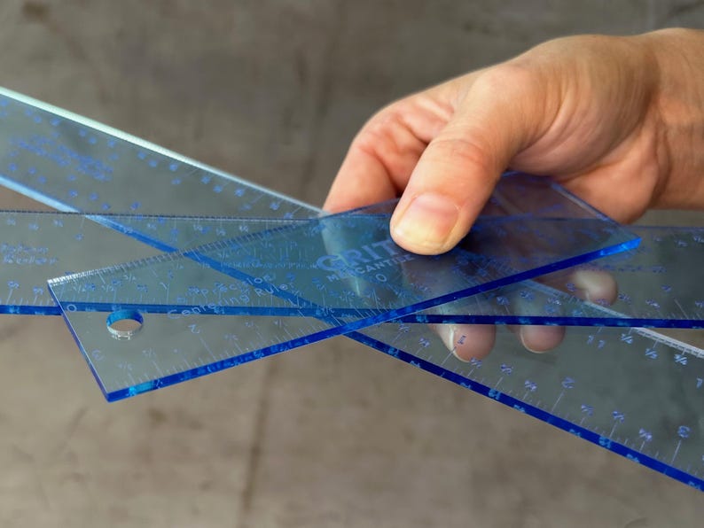 May include: Two blue plastic rulers with a clear background. The rulers are marked with inches and fractions of an inch. The top ruler has the text "GRITT" and "MENSANTILE" printed on it.