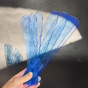 May include: A hand holds a fan of translucent blue plastic tools. Each tool is labeled with the word "GRIT" and has measurement markings and hole patterns. The tools are designed for spacing and are likely used in crafting or woodworking.