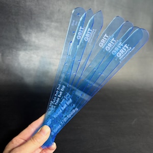 May include: Six blue plastic belt end templates with the text "GRIT MERCANTILE gritmercantile.com" printed on them. The templates are labeled with "Skive This", "High Point Belt End", "Click Point Belt End", "Round Belt End", "Holes 7/8 Spacing", and "60°"